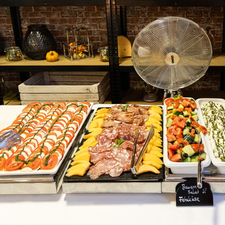 Kaltes Vorspeisenbuffet mit Tomate-Mozzarella, Schinken mit Melone, Bauernsalat, mariniertem Fetakäse und Antipasti vor einem Tischventilator.